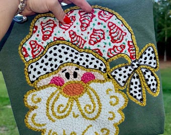 Peut inclure: Un article en tissu vert avec un motif de Père Noël. Le Père Noël a une barbe blanche, un nez orange et des joues roses. Son chapeau est rouge et blanc avec des motifs de cannes en bonbon, et un nœud à pois noir et blanc est attaché.