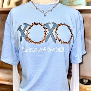 May include: Light blue t-shirt with the text "XOXO" in a stylized design featuring nails and a crown of thorns. The phrase "the original love letter" is printed below the design. A necklace is visible at the neckline.