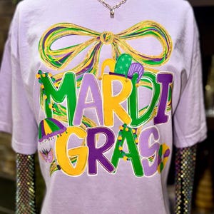 May include: A light purple t-shirt with a large, colorful "Mardi Gras" graphic. The design features a bow, jester hats, and the text in green, yellow, and purple. A gold chain necklace is visible at the neckline.