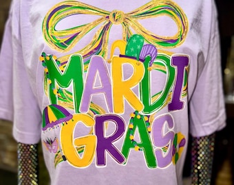 May include: A light purple t-shirt with a large, colorful "Mardi Gras" graphic. The design features a bow, jester hats, and the text in green, yellow, and purple. A gold chain necklace is visible at the neckline.
