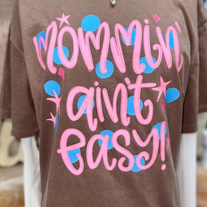 May include: Brown t-shirt with the phrase "Mommin ain't easy!" in pink script, accented with blue circles and stars. The shirt is worn over a white long-sleeved shirt and accessorized with gold necklaces.