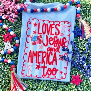 Può includere: Una maglietta blu chiaro con una grafica di bandiera americana rossa, bianca e blu e il testo "LOVES Jesus and AMERICA too" in lettere rosse.