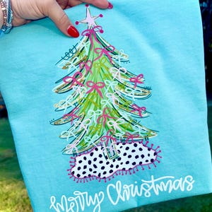 May include: A light blue Christmas-themed sweatshirt featuring a whimsical Christmas tree design. The tree is decorated with pink bows, gold accents, and a star on top. The words "Merry Christmas" are written in white script at the bottom.