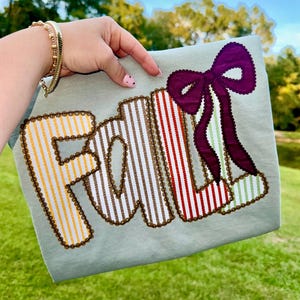 May include: A light green fabric item with the word "FALL" embroidered in colorful striped patterns. A large purple bow is attached to the right side. The letters and bow are outlined with a brown beaded trim. The background is a green lawn and trees.