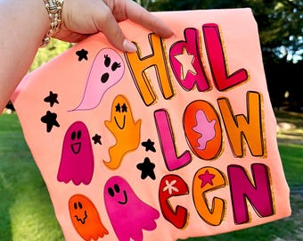 May include: Peach-coloured Halloween-themed t-shirt with the word "HALLOWEEN" in colourful, outlined letters. The shirt features cartoon ghosts and stars in various colours, including pink, orange, and black. The shirt is held up against a green background.