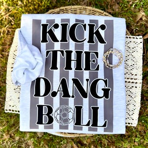 May include: A light gray t-shirt with vertical gray stripes and the text "KICK THE DANG BALL" in bold black letters. A soccer ball graphic is at the bottom. A rolled-up gray sleeve is visible on the left. A bracelet is on the right.