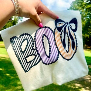 May include: Cream-colored t-shirt with the word "BOO" in a Halloween-themed design. The "B" is black with vertical stripes, the "O" is purple with vertical stripes, and the last "O" is orange with vertical stripes. A black bow is at the top.