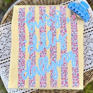 May include: A yellow tote bag with vertical floral stripes and the words "busy lilly dallying" in light blue script. A light blue floral hair clip rests on the top right corner.
