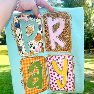 May include: A light blue t-shirt with the word "PRAY" in a colorful, block letter design. The letters are in individual squares with different patterns, including floral, leopard print, plaid, and polka dots. The shirt is held up by a person.