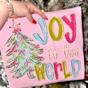 May include: Pink t-shirt with a Christmas tree graphic and the words "JOY to the WORLD" in colorful letters. The tree is decorated with bows and a star. The shirt is a festive holiday item.