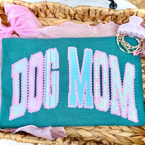 May include: Teal t-shirt with the words "DOG MOM" in a decorative font. The letters are outlined in pink and filled with blue polka dots and floral patterns. A pink bow and bracelets are in the background.