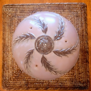 May include: A vintage ceiling light fixture with a pale pink glass shade featuring a silver leaf design. The shade is mounted on a square woven wicker base. The center of the shade has a decorative swirl design.