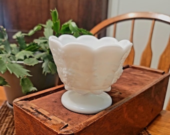 May include: A white, vintage-style glass bowl with a scalloped rim and a pedestal base. The bowl features a grape design and sits on a wooden box. The bowl is likely used for serving or display.
