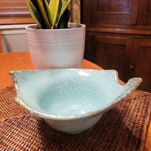 May include: A light turquoise ceramic bowl with a speckled finish and metallic gold-toned edges. The bowl features an irregular, organic shape with pointed, flared sides that curve upward, creating a distinct, wide-rimmed silhouette.