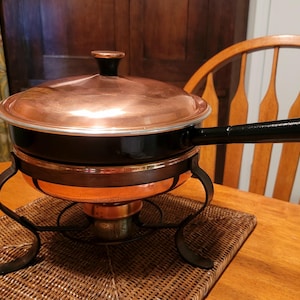 May include: Copper-colored fondue pot with a black handle and lid. The pot sits on a black metal stand with curved legs. The pot is on a woven placemat on a wooden table.