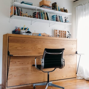 May include: A wooden desk with a fold-down bed, a black office chair, and white shelves with books and decorative items.