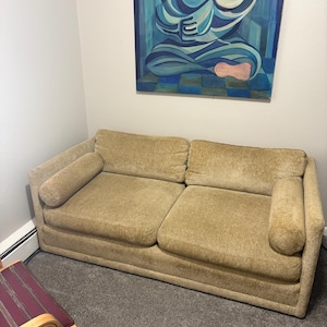 May include: A two-seater sofa in a light brown, textured fabric. The sofa has two back cushions and two cylindrical armrests. A painting with blue tones hangs on the wall above the sofa. A chair with a striped seat is in the foreground.