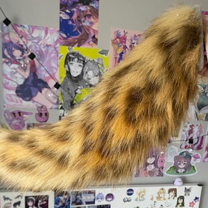 May include: A faux fur tail with a spotted pattern in shades of brown and tan. The tail is held up against a wall decorated with anime-style art prints and stickers. The tail appears to be soft and fluffy.