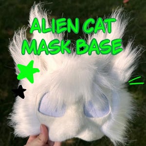 Peut inclure: Une base de masque de chat extraterrestre en fausse fourrure blanche avec de grandes ouvertures pour les yeux et des oreilles pointues. Le texte ALIEN CAT MASK BASE est imprimé en vert, avec une étoile verte et une petite étoile noire sur le côté.