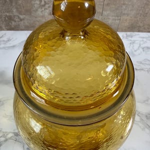 May include: A translucent amber-colored glass jar with a matching lid. The jar and lid feature a textured, hammered surface pattern. The lid is topped with a smooth, spherical glass knob handle, creating a rounded silhouette for the container.