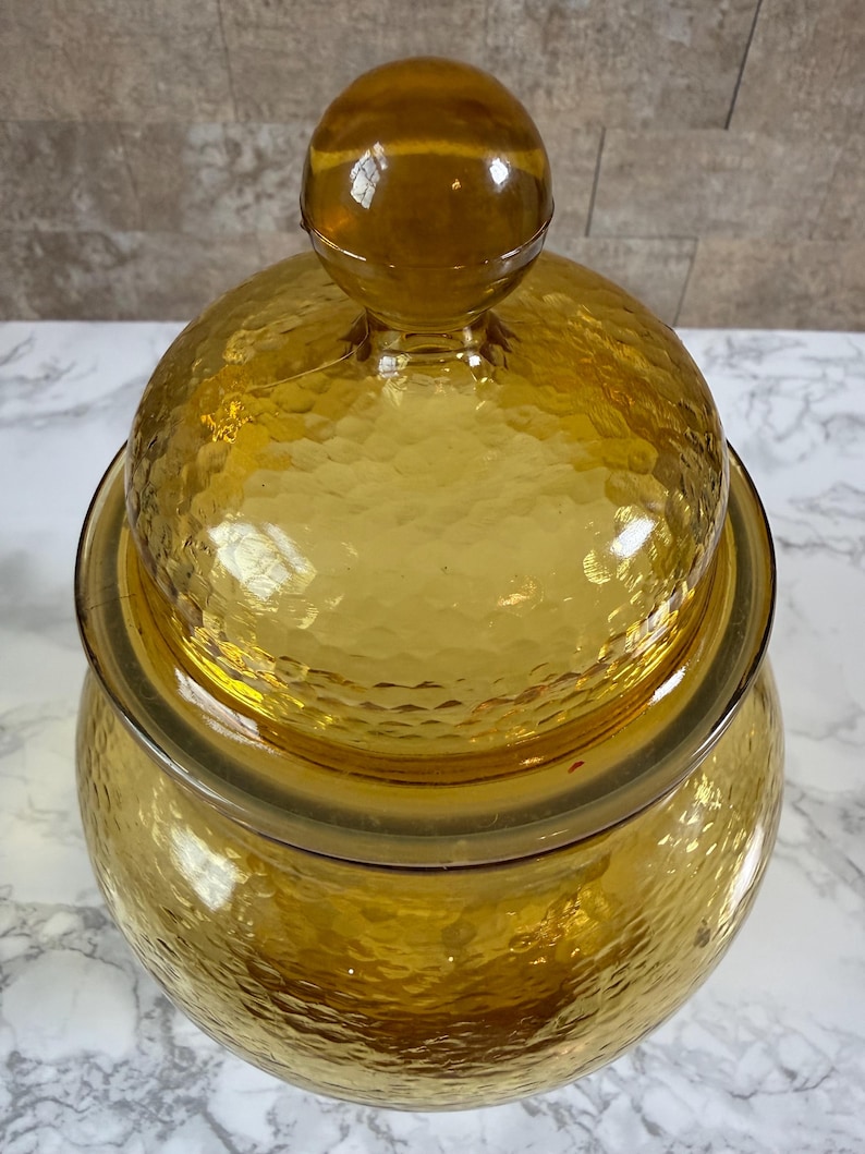 May include: A translucent amber-colored glass jar with a matching lid. The jar and lid feature a textured, hammered surface pattern. The lid is topped with a smooth, spherical glass knob handle, creating a rounded silhouette for the container.