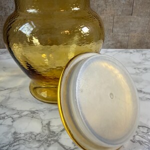 May include: A large, amber-colored glass jar with a textured, hammered surface and a wide, rounded body. The jar features a matching amber glass lid with a clear, circular plastic seal on the underside, designed to fit securely into the jar opening.