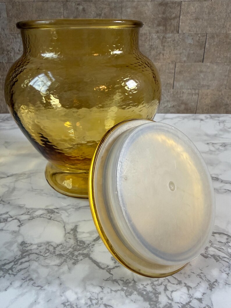 May include: A large, amber-colored glass jar with a textured, hammered surface and a wide, rounded body. The jar features a matching amber glass lid with a clear, circular plastic seal on the underside, designed to fit securely into the jar opening.