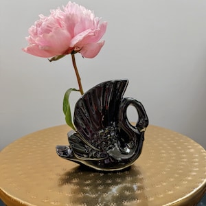 May include: A black ceramic swan vase with a pink peony flower. The swan vase has a glossy finish and a curved neck. The peony is a light pink color and has many petals. The vase is on a gold-colored table.