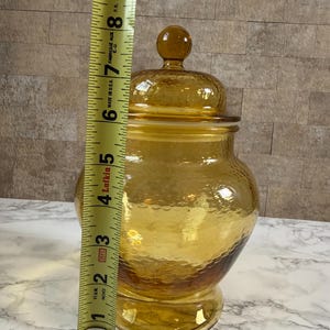 May include: An amber-colored glass jar with a matching lid and a textured, dimpled surface. A yellow measuring tape is positioned vertically against the jar, indicating a total height of approximately 9 inches from the base to the lid finial.