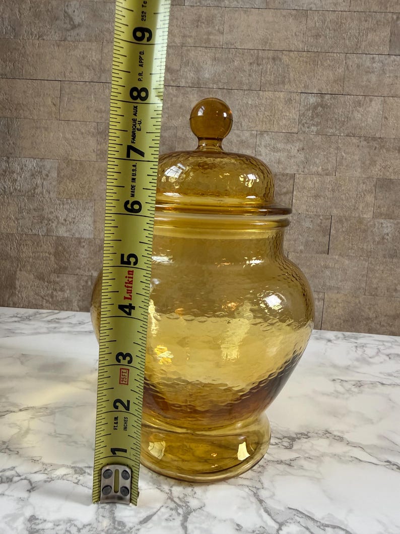May include: An amber-colored glass jar with a matching lid and a textured, dimpled surface. A yellow measuring tape is positioned vertically against the jar, indicating a total height of approximately 9 inches from the base to the lid finial.