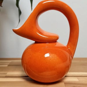 May include: A vibrant orange ceramic pitcher with a unique, curved handle and spout. The pitcher has a rounded base and a glossy finish. The design is modern and sculptural, suitable for decorative use. The pitcher is approximately 10 inches tall.