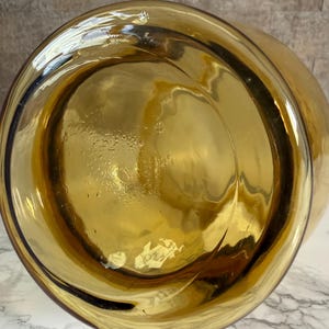 May include: A circular amber-colored glass vessel base viewed from directly underneath. The glass is translucent with a smooth, reflective surface and subtle internal ripples. A small, white, rectangular adhesive label is attached to the bottom.