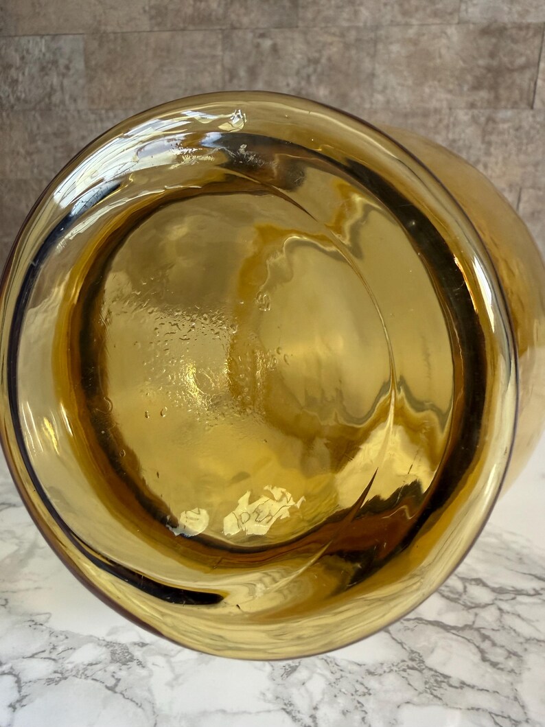 May include: A circular amber-colored glass vessel base viewed from directly underneath. The glass is translucent with a smooth, reflective surface and subtle internal ripples. A small, white, rectangular adhesive label is attached to the bottom.