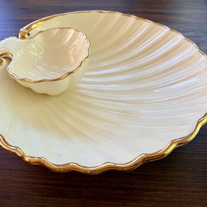 May include: A cream-colored ceramic serving dish shaped like a large scallop shell with a smaller, matching shell-shaped bowl attached to one side. Both pieces feature a scalloped edge and are finished with a metallic gold trim along the rims.
