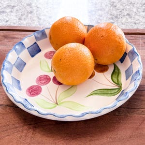 May include: Three oranges rest on a decorative ceramic plate. The plate features a scalloped edge with a blue and white checkered pattern, and a painted cherry and leaf design. The oranges are bright orange and have a textured peel.