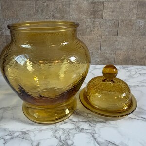May include: A large, amber-colored glass jar with a wide mouth and a matching removable lid. The glass features a distinct hammered texture throughout the surface, including the rounded lid with a small knob handle and the wide, stable base.