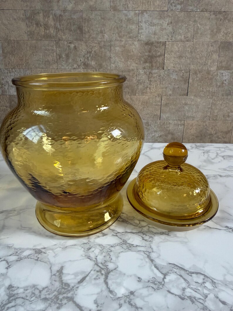 May include: A large, amber-colored glass jar with a wide mouth and a matching removable lid. The glass features a distinct hammered texture throughout the surface, including the rounded lid with a small knob handle and the wide, stable base.