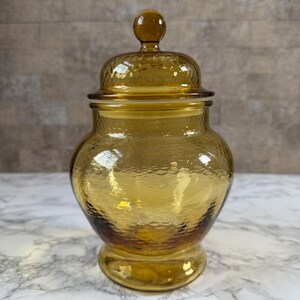 May include: A translucent amber-colored glass jar with a rounded body and a matching lid. The glass features a textured, hammered surface throughout the exterior. The lid is topped with a small, spherical knob handle for easy removal.