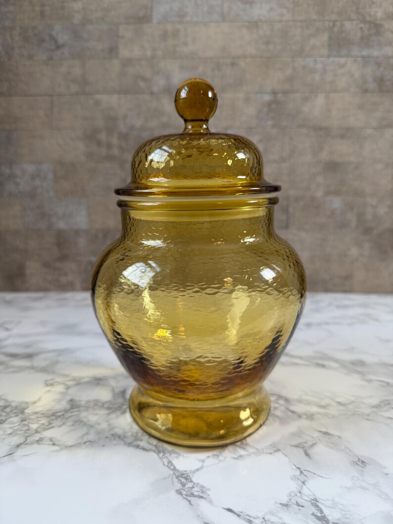 May include: A translucent amber-colored glass jar with a rounded body and a matching lid. The glass features a textured, hammered surface throughout the exterior. The lid is topped with a small, spherical knob handle for easy removal.