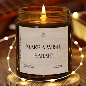 Peut inclure: Une bougie en verre brun avec un couvercle en bois et une étiquette blanche qui dit "Make a Wish, Sarah!" La bougie est allumée et entourée de lumières féériques blanches.