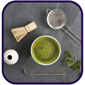 May include: A white ceramic bowl filled with frothy green matcha tea, a bamboo whisk, a metal tea strainer, a wooden tea scoop, and a sprig of green leaves on a gray stone surface.