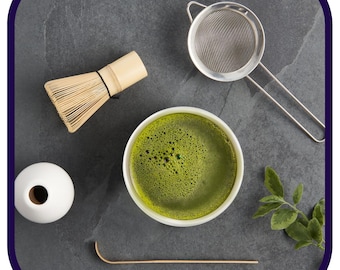 May include: A white ceramic bowl filled with frothy green matcha tea, a bamboo whisk, a metal tea strainer, a wooden tea scoop, and a sprig of green leaves on a gray stone surface.