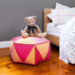 Könnte beinhalten: Ein gepolsterter Hocker mit rosafarbenem, orangefarbenem und gelbem Muster, auf dem ein Teddybär sitzt. Der Hocker steht in einem Schlafzimmer mit einem Bett im Hintergrund.
