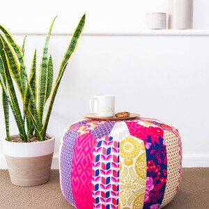 May include: A colorful patchwork fabric pouf ottoman with a white mug and a cookie on a wooden tray on top. The pouf is sitting on a brown carpet.