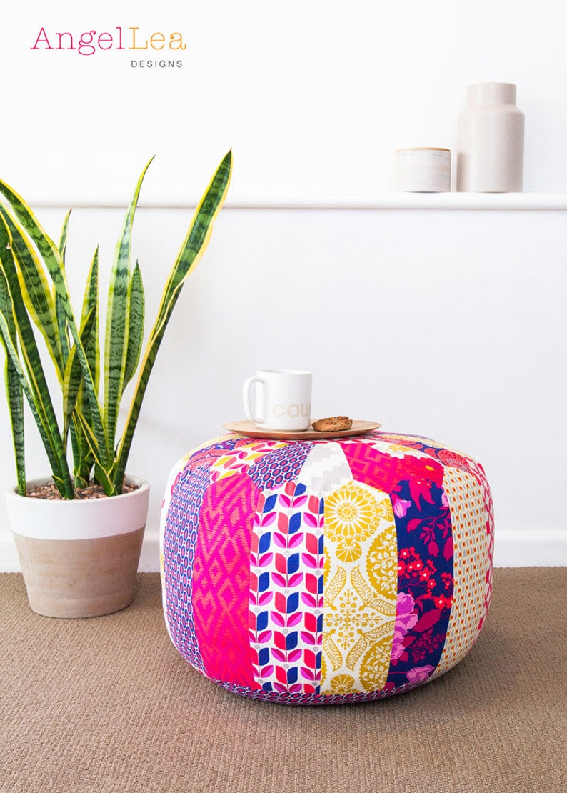 May include: A colorful patchwork fabric pouf ottoman with a white mug and a cookie on a wooden tray on top. The pouf is sitting on a brown carpet.