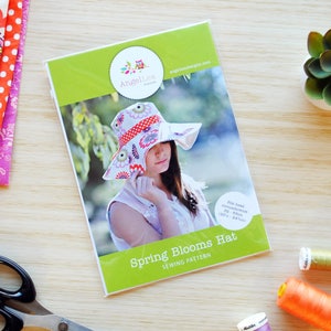 May include: A green and white sewing pattern for a Spring Blooms Hat. The pattern features a photo of a woman wearing a floral hat. The text "Spring Blooms Hat" and "Sewing Pattern" is printed on the pattern.
