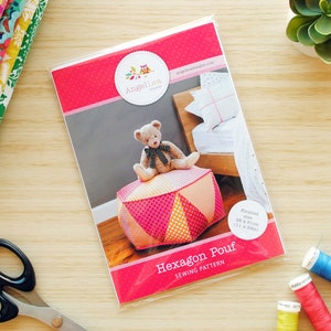 Könnte beinhalten: Ein Nähmuster für einen rosa und gelben sechseckigen Hocker mit einem Teddybären darauf. Das Muster enthält Anweisungen für die Herstellung eines Hockers mit einer Größe von 71 cm x 71 cm.