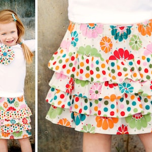 May include: A white long-sleeved shirt with a colourful flower embellishment on the front, paired with a multi-layered skirt featuring a white background with colourful floral and polka dot patterns.