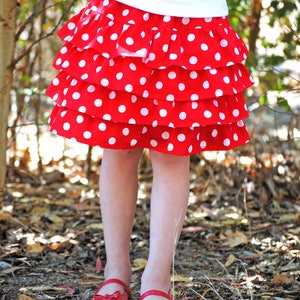 May include: A red and white polka dot skirt with multiple ruffles. The skirt is worn by a child and is photographed against a background of brown leaves.