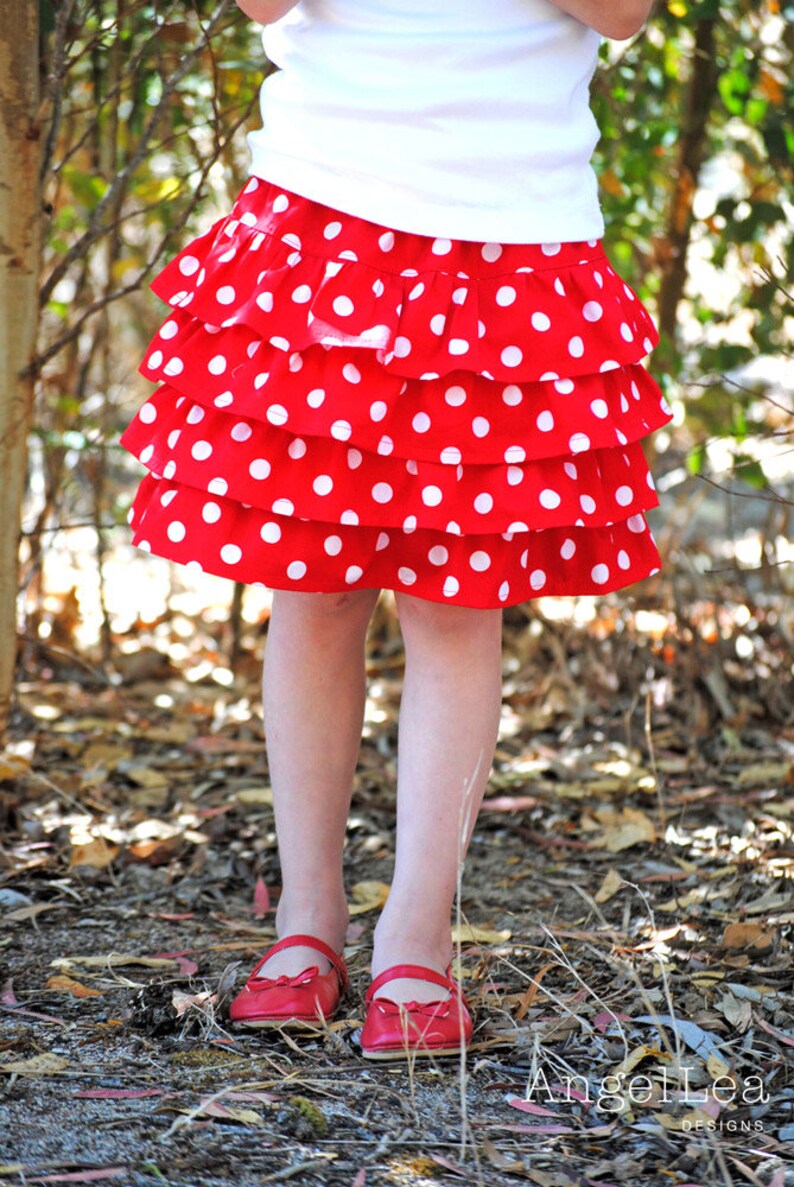 May include: A red and white polka dot skirt with multiple ruffles. The skirt is worn by a child and is photographed against a background of brown leaves.